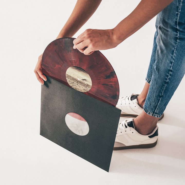 A person wearing white sneakers and blue jeans is holding a red marbled vinyl record, partially removing it from a Groove Guardian Black Poly-Lined Paper Record Sleeve with a round cutout on a white background, ensuring optimal vinyl protection.