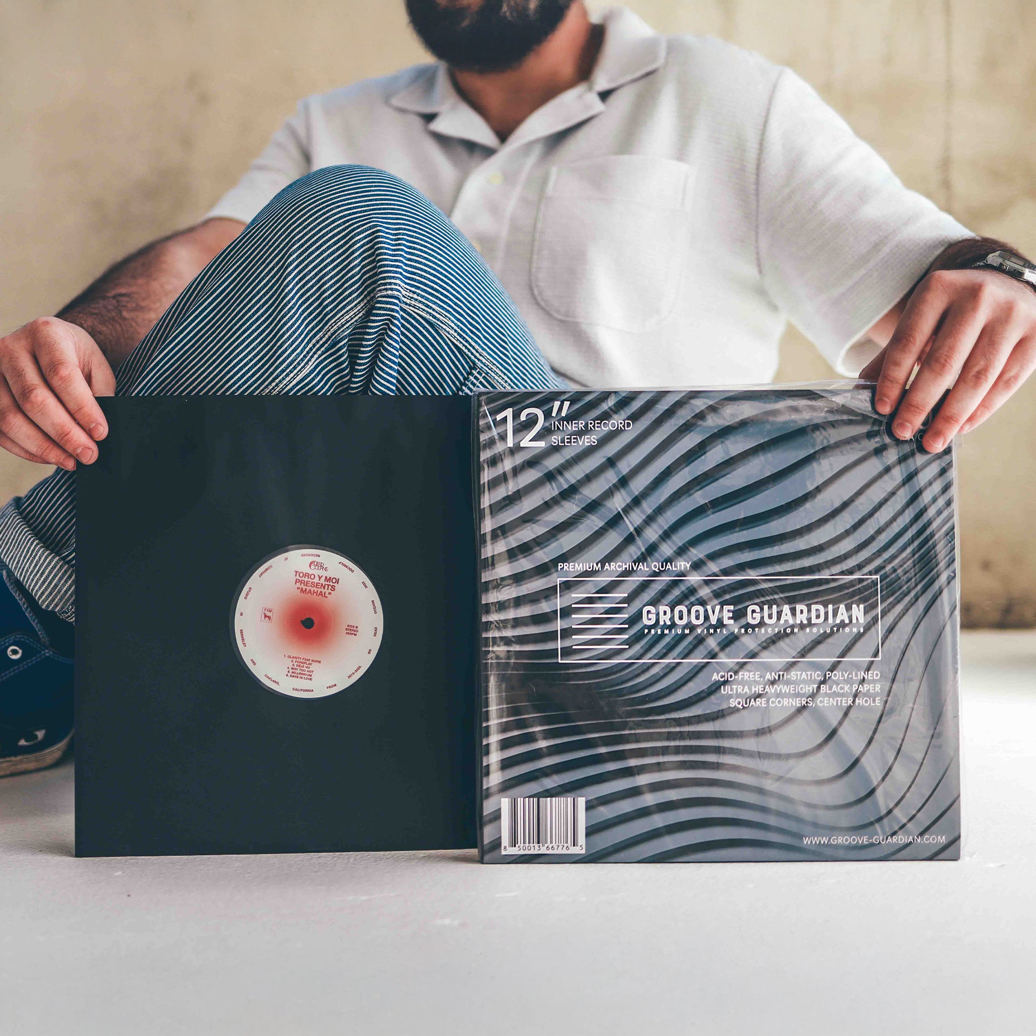 A person in a white shirt and striped pants sits cross-legged, holding a vinyl record next to its Groove Guardian sleeve—featuring abstract black-and-white lines designed for premium protection. Only their lower face and hands are visible, while nearby, Groove Guardian Black Poly-Lined Paper Record Sleeves for 12” records hint at a careful collector’s touch.