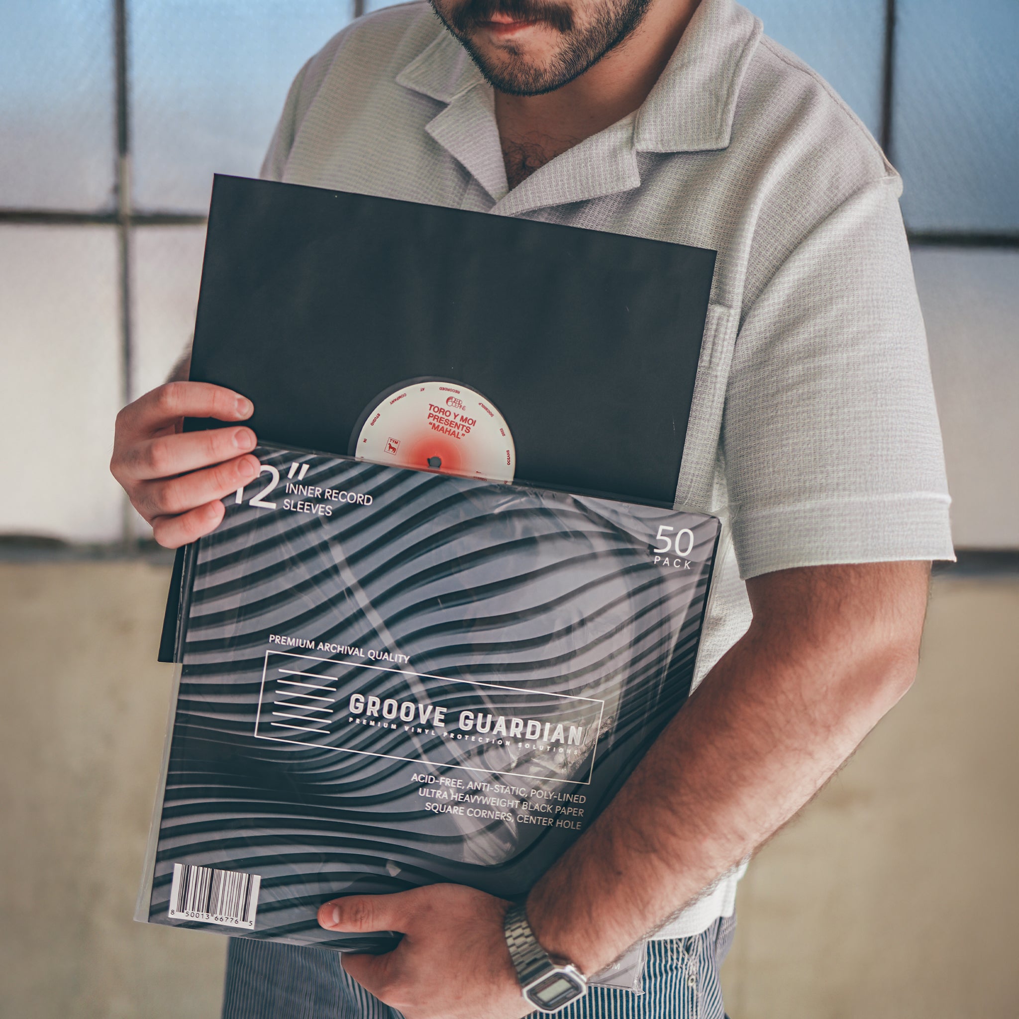 A person in a light shirt stands indoors by a window, holding a vinyl record and a pack of Groove Guardian Black Poly-Lined Paper Record Sleeves in front of their chest—demonstrating their dedication to premium vinyl protection.