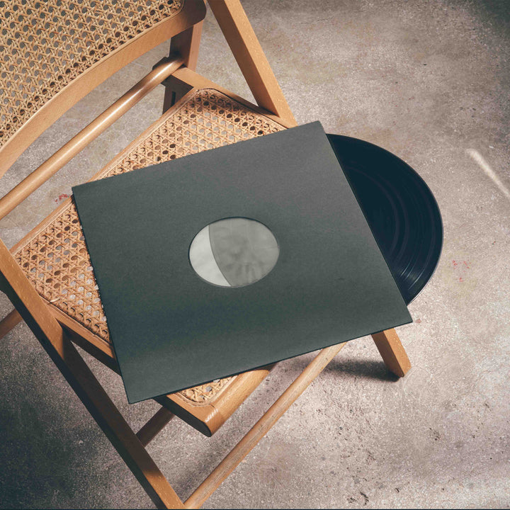A black vinyl record, securely placed inside a Groove Guardian Black Poly-Lined Paper Record Sleeve for premium protection, rests on a wooden chair with a woven cane seat atop a concrete floor.