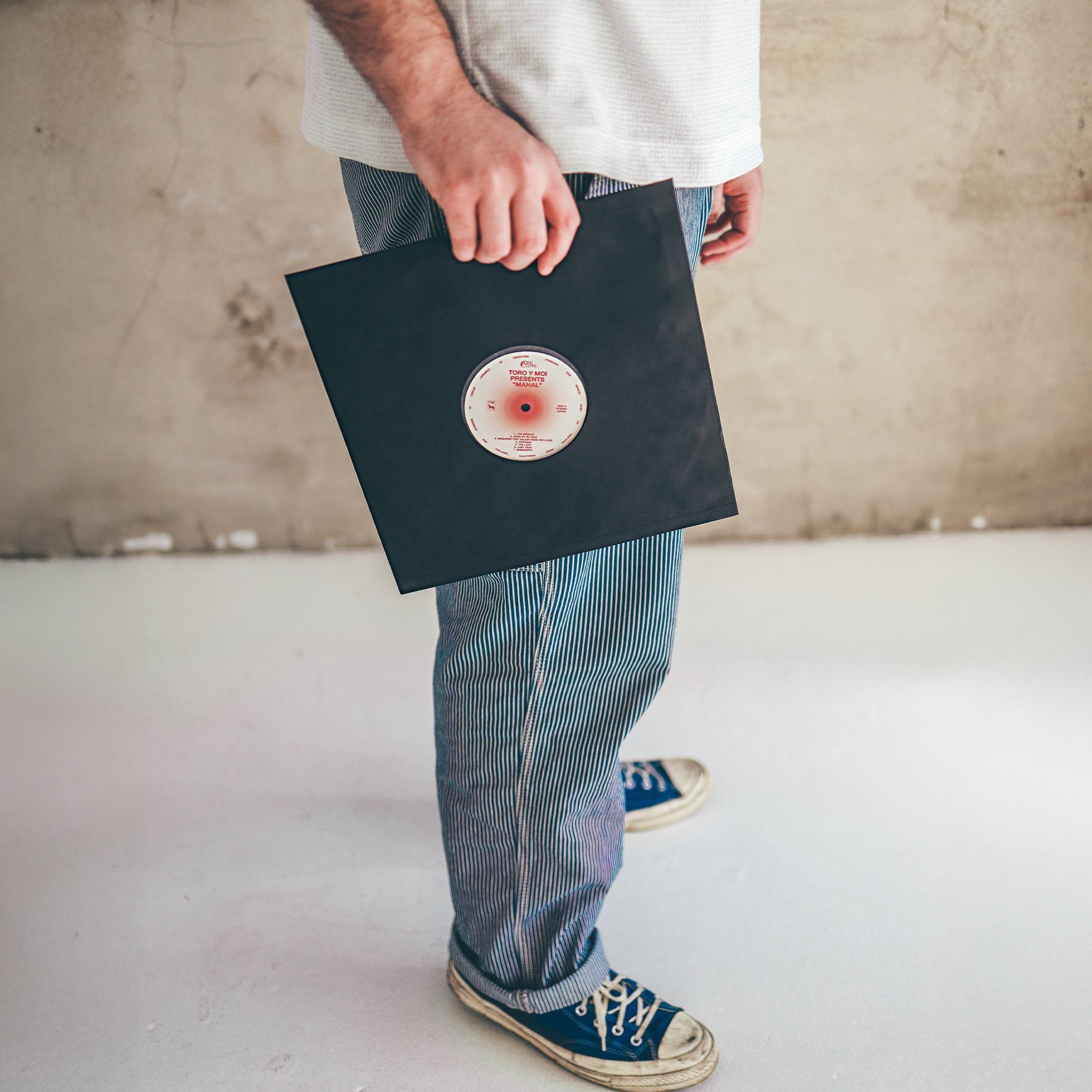 A person in blue striped pants and blue sneakers holds a vinyl record in a Groove Guardian Black Poly-Lined Paper Record Sleeve by their side, standing against a plain wall for added vinyl record protection.