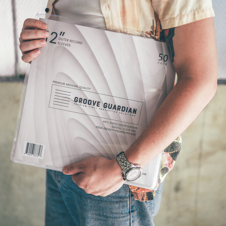 A person in a short-sleeve shirt holds a pack of Groove Guardian Premium 12" LP Outer Record Sleeves. The pack contains 50 high-quality sleeves for 12" vinyl records and features a minimalist design. The person, wearing a watch, stands near a fence.