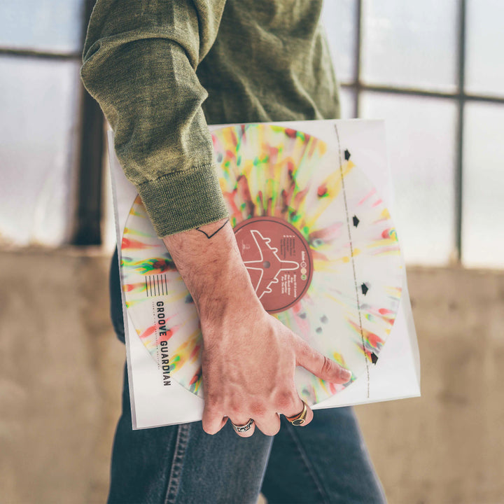 A person in a green sweater holds a colorful, splattered vinyl record protected in a Groove Guardian Premium 12" LP Inner Record Sleeve, standing near large windows with only their torso and hand visible.