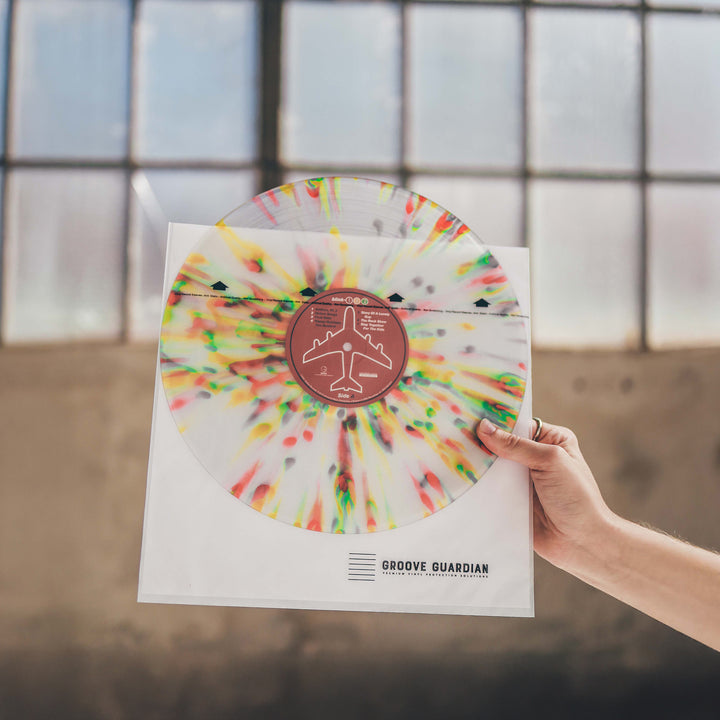 A hand holds a vibrant splatter-patterned vinyl record partially enclosed in a clear sleeve labeled "Groove Guardian," featuring archival-quality protection with Groove Guardian Premium 12" LP Inner Record Sleeves—all showcased against industrial-style windows.
