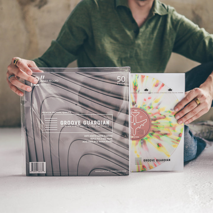 A person in a green shirt sits on the floor, holding vinyl record sleeves and a colorful splatter-patterned record. The sleeve is labeled Groove Guardian Premium 12" LP Inner Record Sleeves, featuring archival-quality protection and essential information about its anti-static properties.