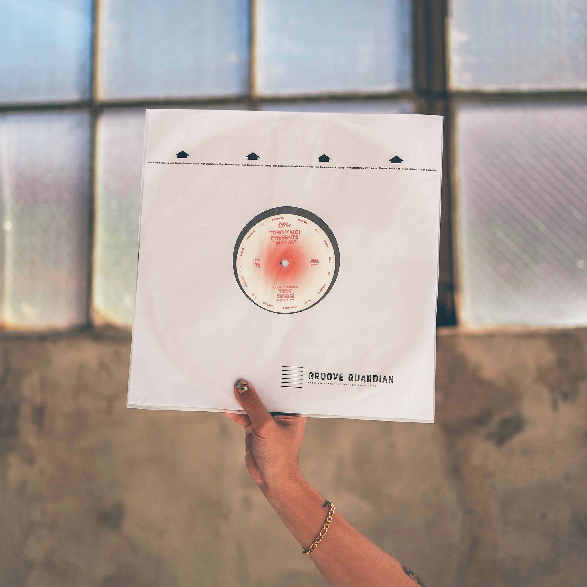A hand with a bracelet holds up a vinyl record in an Ultra-Premium 12" LP Inner Record Sleeve from Groove Guardian, designed to protect 12-inch records, with a blurred window and textured wall in the background.