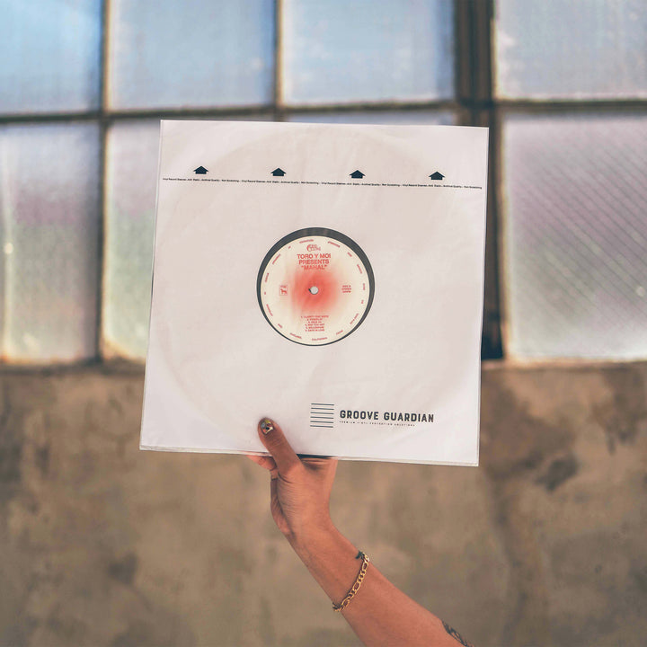A hand with a bracelet holds up a vinyl record in an Ultra-Premium 12" LP Inner Record Sleeve from Groove Guardian, designed to protect 12-inch records, with a blurred window and textured wall in the background.