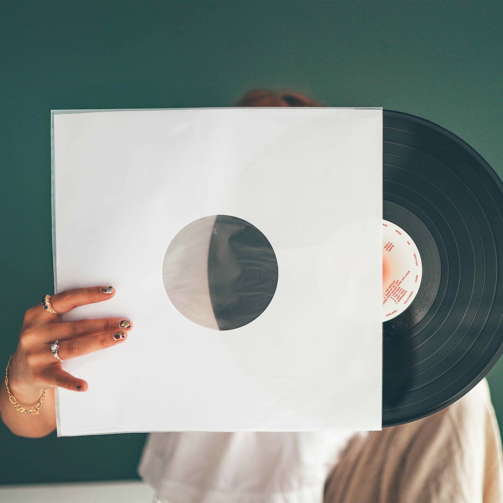 A person holds a Groove Guardian Ultra-Premium 12" LP Inner Record Sleeve, featuring a circular window and a white design, partially obscuring their face against a dark green background.